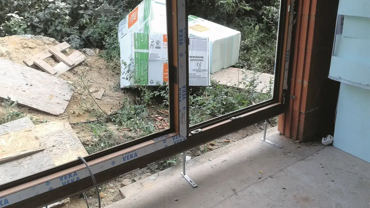Próg drzwi tarasowych w trakcie montażu, przygotowany do ocieplenia progu. W tle materiały budowlane i zieleń.