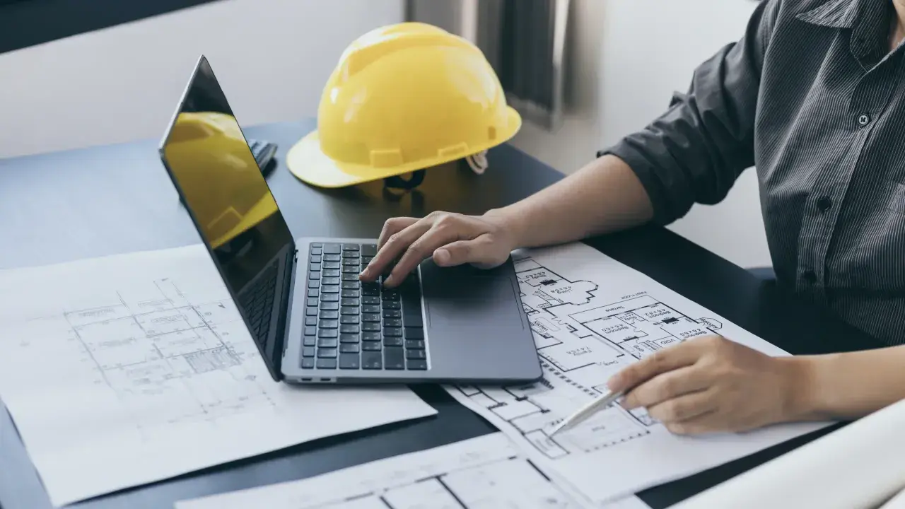 Architekt analizuje plany, porównując powierzchnię zabudowy z powierzchnią użytkową. Na biurku leży żółty kask.