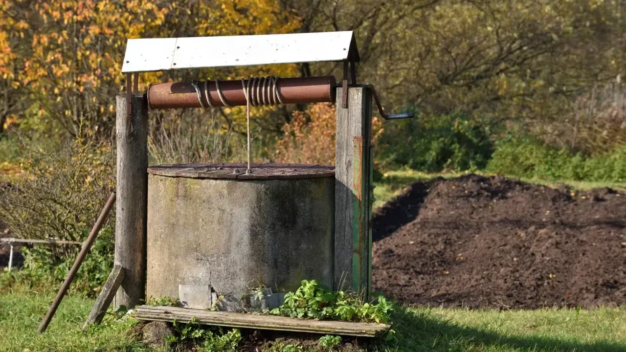 Stary, betonowy krąg studni kręgowej z drewnianą konstrukcją i korbą. W tle jesienne drzewa i kopiec ziemi.