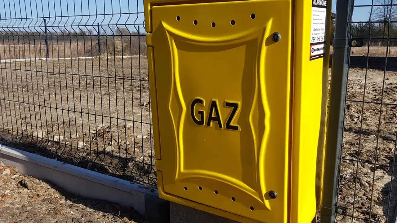Żółta skrzynka gazowa z napisem "GAZ" przy ogrodzeniu. To element przyłącza gazu od skrzynki do domu.