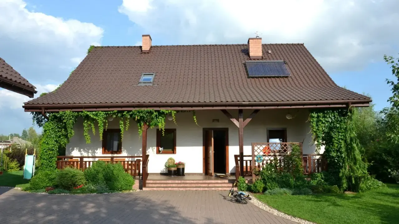 Urokliwy wiejski dom z werandą, otoczony zielenią. Na dachu panele słoneczne.