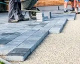 Układanie betonowych płyt na żwirze. Pracownicy tworzą nowoczesny taras, gdzie beton zamiast kostki brukowej stanowi estetyczne i trwałe rozwiązanie.
