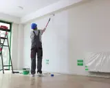 Malowanie ścian w nowym mieszkaniu. Pracownik w kasku i ogrodniczkach maluje wałkiem białą farbą. Obok drabina, wiadro z farbą i folia ochronna. Zastanawiasz się, ile za m2 malowania?
