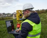 Geodeta w kamizelce odblaskowej i kasku pracuje z laptopem i tachimetrem w terenie. Zastanawiasz się, ile bierze geodeta za wytyczenie działki?