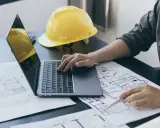 Architekt analizuje plany, porównując powierzchnię zabudowy z powierzchnią użytkową. Na biurku leży żółty kask.