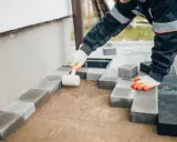 Ręce w rękawiczkach układają kostkę brukową, tworząc opaskę z kostki brukowej przy ścianie domu.
