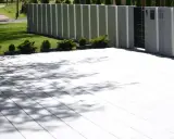 Nowoczesny taras z płyt betonowych, otoczony zielenią i nowoczesnym ogrodzeniem. Cienie drzew tańczą na jasnej nawierzchni.
