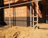 Budowa domu: widoczny fragment ściany z cegły, ciemny cokół co to jest, rusztowanie i piasek na placu budowy.