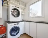 Nowoczesna pralnia w domu z pralką i suszarką ustawionymi jedna na drugiej. Obok okno i białe szafki.