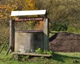 Stary, betonowy krąg studni kręgowej z drewnianą konstrukcją i korbą. W tle jesienne drzewa i kopiec ziemi.