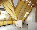 Widok na poddasze w trakcie prac. Drewniana konstrukcja dachu, okna dachowe i żółta wełna mineralna świadczą o postępie prac nad adaptacją poddasza. Projekt w toku.