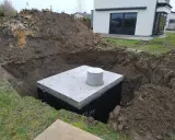 Na działce trwa montaż szamba. Betonowy zbiornik jest już częściowo zakopany w wykopie, otoczony ziemią.
