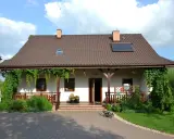 Urokliwy wiejski dom z werandą, otoczony zielenią. Na dachu panele słoneczne.