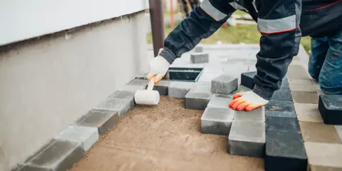 Ręce w rękawiczkach układają kostkę brukową, tworząc opaskę z kostki brukowej przy ścianie domu.
