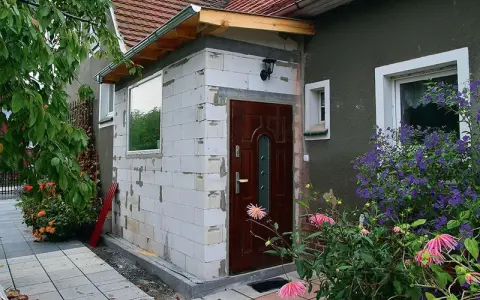 Rozbudowa domu na zgłoszenie w toku. Nowa ściana z bloczków, drzwi i okno czekają na wykończenie. Wokół kwitną kwiaty.
