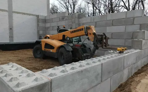 Żółty ładowacz teleskopowy Liebherr pracuje przy budowie muru oporowego z betonowych bloków.