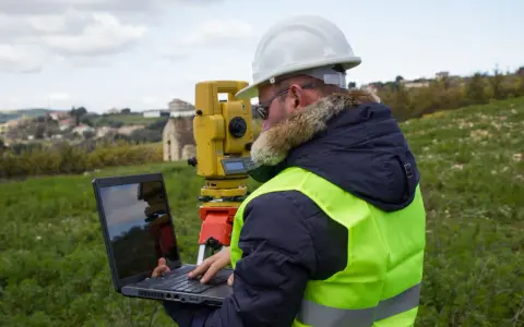 Geodeta w kamizelce odblaskowej i kasku pracuje z laptopem i tachimetrem w terenie. Zastanawiasz się, ile bierze geodeta za wytyczenie działki?