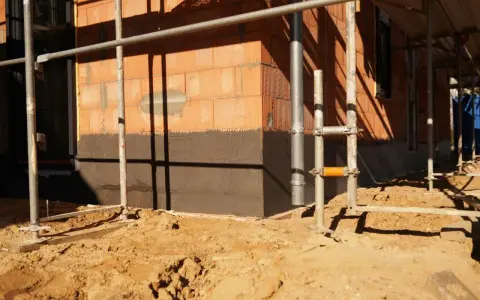 Budowa domu: widoczny fragment ściany z cegły, ciemny cokół co to jest, rusztowanie i piasek na placu budowy.
