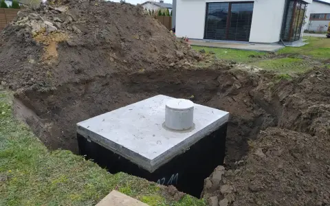 Na działce trwa montaż szamba. Betonowy zbiornik jest już częściowo zakopany w wykopie, otoczony ziemią.