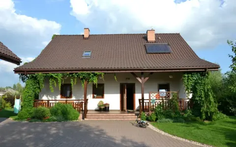 Urokliwy wiejski dom z werandą, otoczony zielenią. Na dachu panele słoneczne.