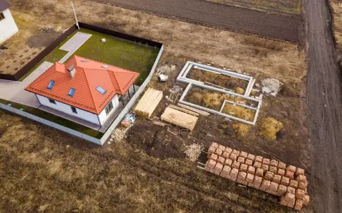 Budowa domu na działce rolnej. Widać fundamenty, sterty drewna i cegieł, gotowy dom. To może być początek planowania, jak podzielić działkę rolną.