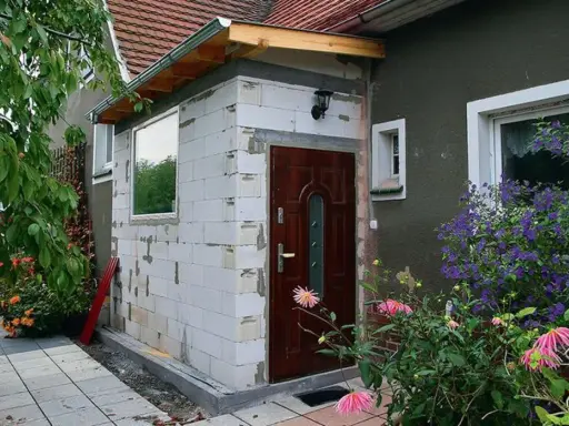 Rozbudowa domu na zgłoszenie w toku. Nowa ściana z bloczków, drzwi i okno czekają na wykończenie. Wokół kwitną kwiaty.