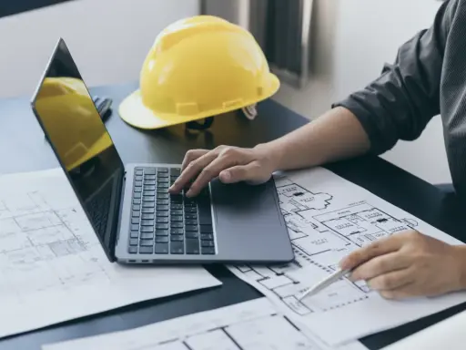 Architekt analizuje plany, porównując powierzchnię zabudowy z powierzchnią użytkową. Na biurku leży żółty kask.
