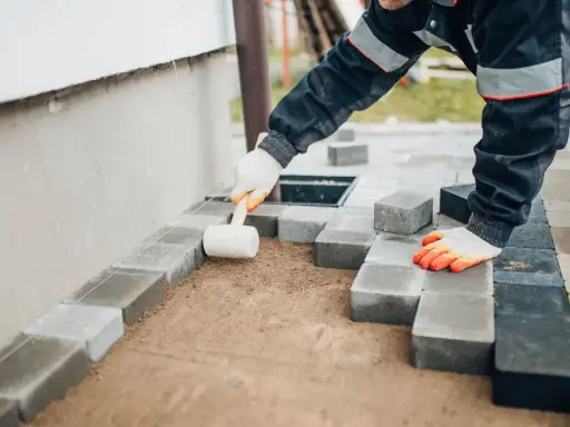 Ręce w rękawiczkach układają kostkę brukową, tworząc opaskę z kostki brukowej przy ścianie domu.