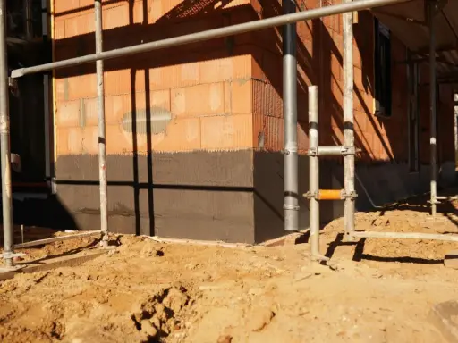 Budowa domu: widoczny fragment ściany z cegły, ciemny cokół co to jest, rusztowanie i piasek na placu budowy.