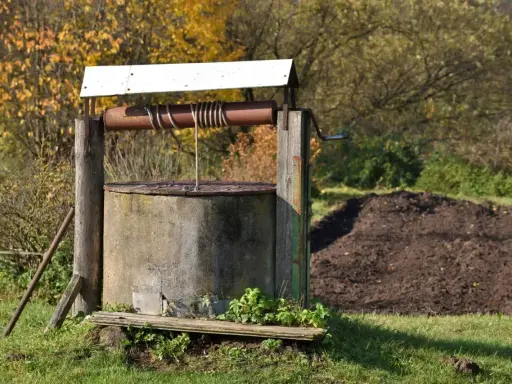 Stary, betonowy krąg studni kręgowej z drewnianą konstrukcją i korbą. W tle jesienne drzewa i kopiec ziemi.