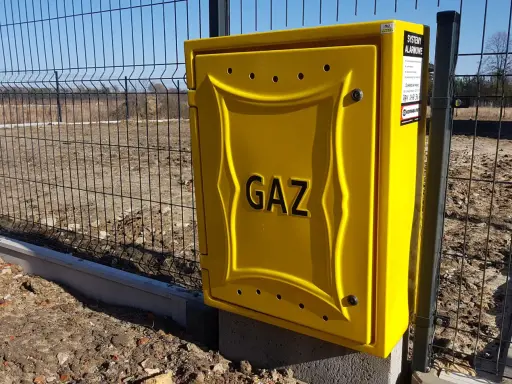 Żółta skrzynka gazowa z napisem "GAZ" przy ogrodzeniu. To element przyłącza gazu od skrzynki do domu.