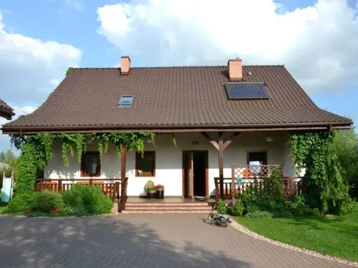 Urokliwy wiejski dom z werandą, otoczony zielenią. Na dachu panele słoneczne.