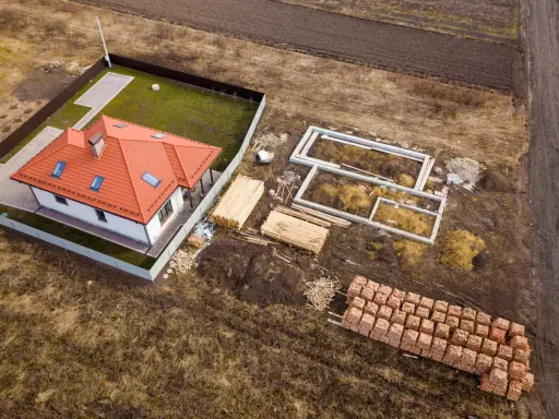 Budowa domu na działce rolnej. Widać fundamenty, sterty drewna i cegieł, gotowy dom. To może być początek planowania, jak podzielić działkę rolną.