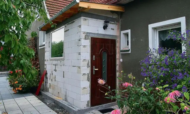 Rozbudowa domu na zgłoszenie w toku. Nowa ściana z bloczków, drzwi i okno czekają na wykończenie. Wokół kwitną kwiaty.