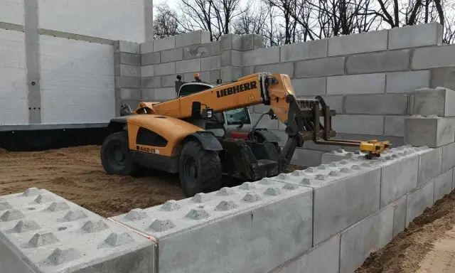 Żółty ładowacz teleskopowy Liebherr pracuje przy budowie muru oporowego z betonowych bloków.