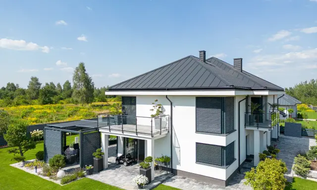 Nowoczesny dom z tarasem na dachu, otoczony zielonym ogrodem i polami kwitnących kwiatów.