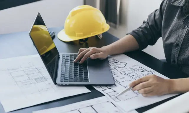 Architekt analizuje plany, porównując powierzchnię zabudowy z powierzchnią użytkową. Na biurku leży żółty kask.