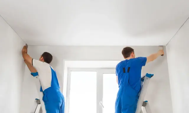 Dwóch fachowców montuje biały sufit napinany. Wzory na suficie są subtelne, tworząc gładką, jednolitą powierzchnię.