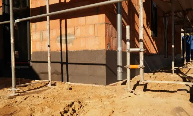Budowa domu: widoczny fragment ściany z cegły, ciemny cokół co to jest, rusztowanie i piasek na placu budowy.