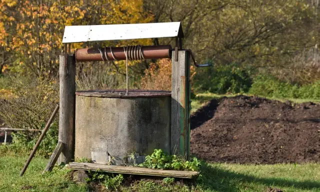 Stary, betonowy krąg studni kręgowej z drewnianą konstrukcją i korbą. W tle jesienne drzewa i kopiec ziemi.