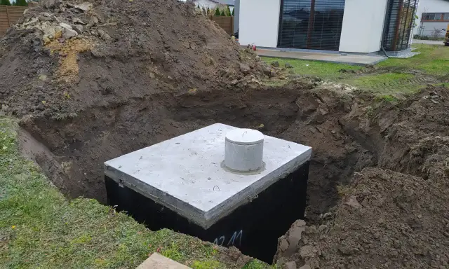 Na działce trwa montaż szamba. Betonowy zbiornik jest już częściowo zakopany w wykopie, otoczony ziemią.