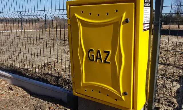 Żółta skrzynka gazowa z napisem "GAZ" przy ogrodzeniu. To element przyłącza gazu od skrzynki do domu.