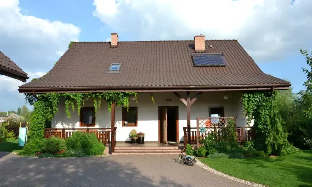 Urokliwy wiejski dom z werandą, otoczony zielenią. Na dachu panele słoneczne.