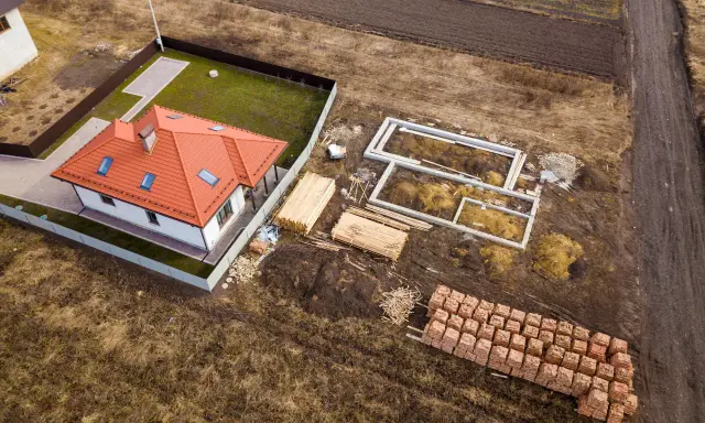 Budowa domu na działce rolnej. Widać fundamenty, sterty drewna i cegieł, gotowy dom. To może być początek planowania, jak podzielić działkę rolną.