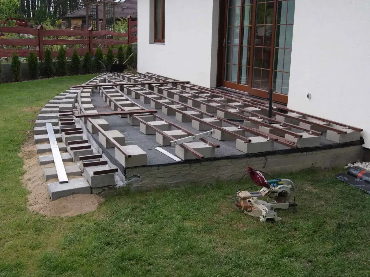 Budowa tarasu z deski kompozytowej. Podbudowa z bloczków betonowych i legary czekają na deski. Obok piła.