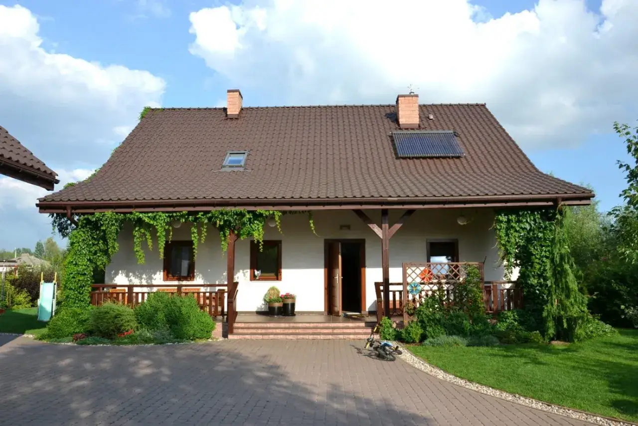 Urokliwy wiejski dom z werandą, otoczony zielenią. Na dachu panele słoneczne.