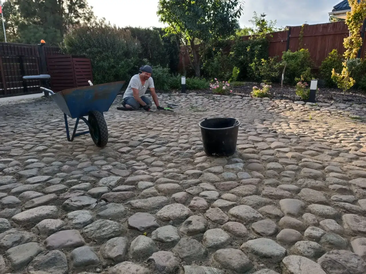 Mężczyzna pracuje przy budowie podjazdu z kamienia. Obok stoi taczka i wiadro.