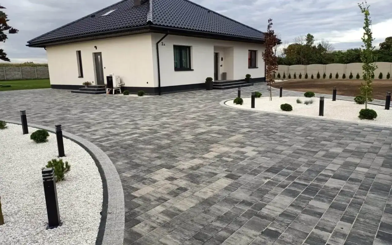 Nowoczesny dom z podjazdem z szarego betonu zamiast kostki brukowej. Oświetlenie i biały żwir tworzą elegancką aranżację.